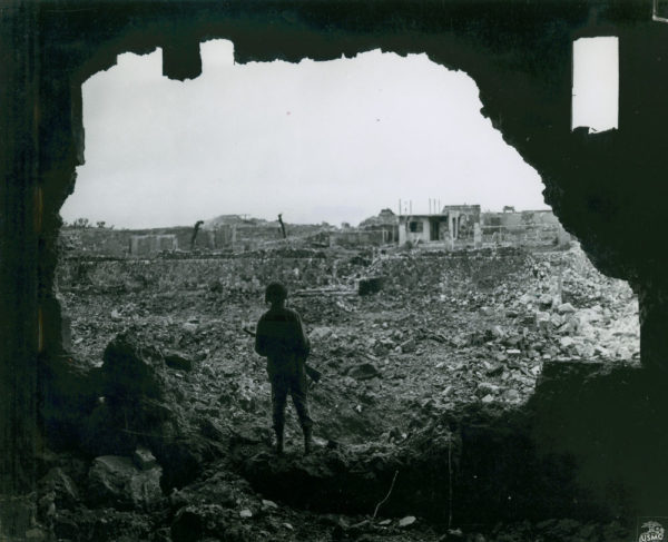 Marine standing within gaping hole of destroyed building