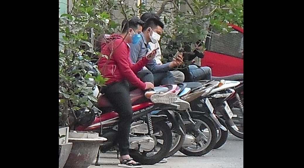 Young men in face masks sitting on their mopeds and looking at their phones