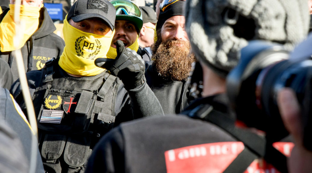 Proud Boys member with yellow bandana over mouth pointing finger at crowd
