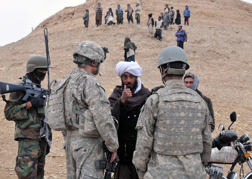 Afghan man flanked by U.S. military personnel, with children on hill in background