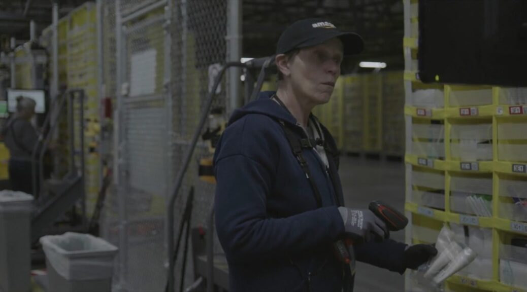 Frances McDormand in an Amazon baseball cap standing in front of pallets in an Amazon warehouse