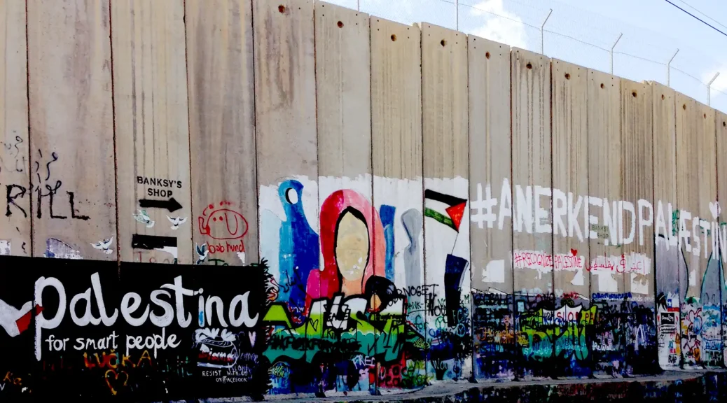 Stone wall with barbed wire on top and colorful graffiti across its breadth